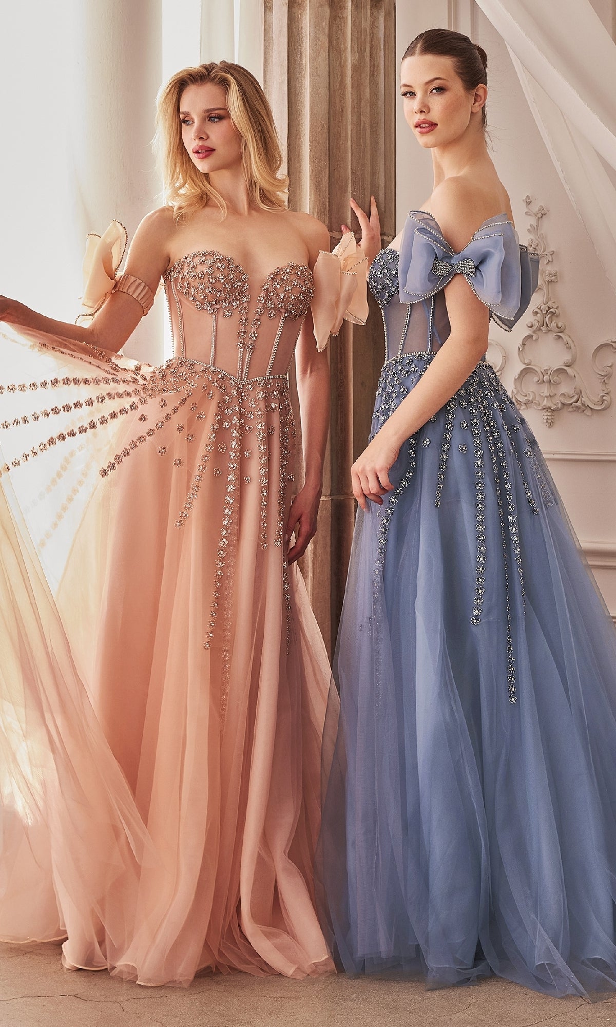Two women stand indoors, each in a strapless Andrea and Leo A1338 prom dress—one peach, one light blue. These elegant, beaded floor-length gowns feature sparkling embellishments, perfect for upscale events.